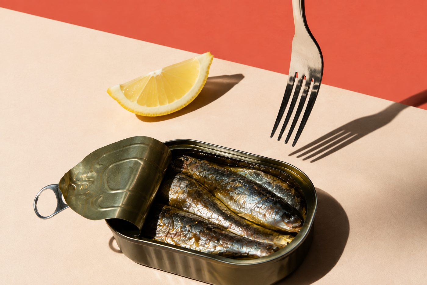 Boîte de sardines ouverte sur fond corail, citron et fourchette